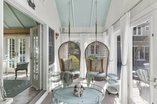 A bright sunroom with hanging chairs, light curtains, and a cozy atmosphere, featuring a glass table and greenery outside.