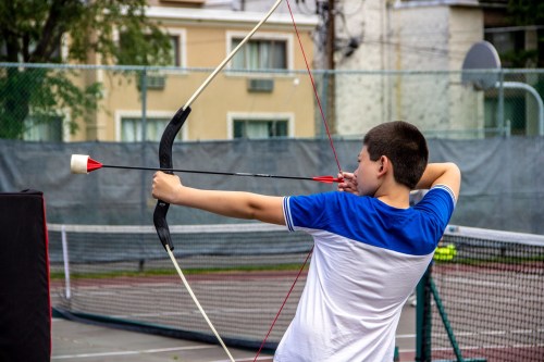 A young boy aims an arrow with a bow on a tennis court, focused and ready to shoot.