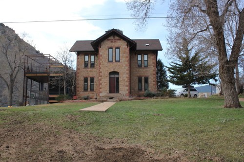 A two-story stone house with a gabled roof, surrounded by grass and trees, set against a rocky hillside.