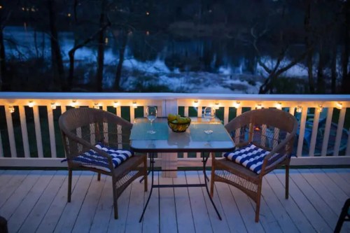 A cozy outdoor seating area with two chairs and a table, overlooking a river at dusk, illuminated by string lights.