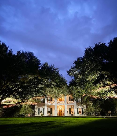 A beautifully lit historic house surrounded by trees, under a dramatic evening sky with scattered clouds.