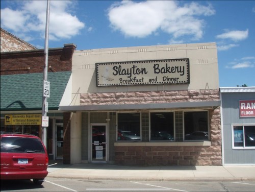 A storefront of Slayton Bakery, featuring a sign that reads "Breakfast and Dinner," with a red car parked in front.