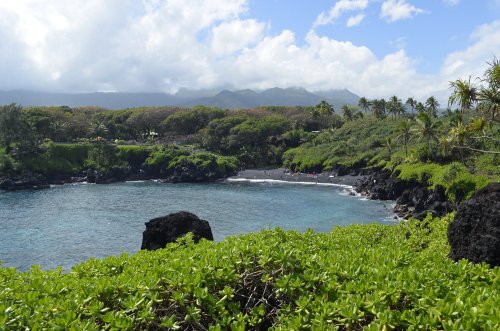A scenic view of a black sand beach surrounded by lush greenery and mountains under a partly cloudy sky.
