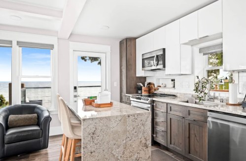 Modern kitchen with white cabinets, granite island, and ocean view through large windows. Cozy seating area included.