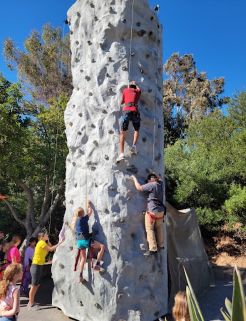 Two climbers ascend a rock wall while a child climbs below, with a group of onlookers in the background.