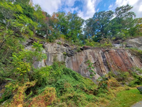 A rocky cliff covered in greenery, with trees above and a blue sky dotted with clouds.