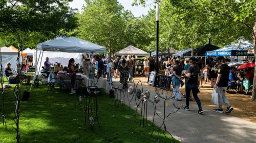 A bustling outdoor market with tents, vendors, and visitors among trees and greenery.