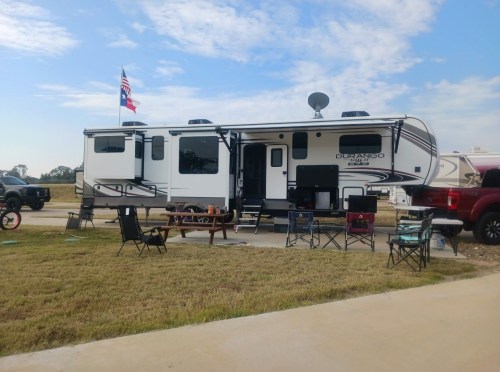 A parked RV with an American and Texas flag, surrounded by chairs and a picnic table on a grassy area.