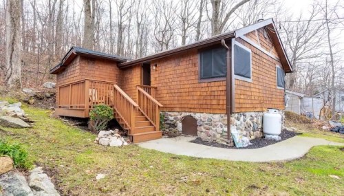 A cozy wooden cabin with a stone foundation, surrounded by trees and a grassy area. Steps lead to the front door.