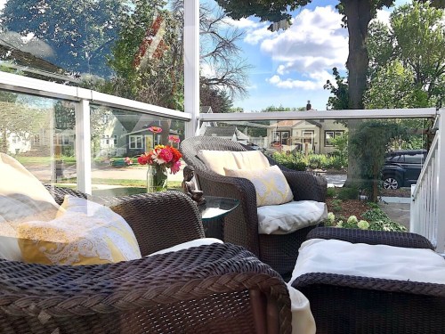 Cozy porch with wicker chairs, decorative pillows, a vase of flowers, and a view of a sunny neighborhood.