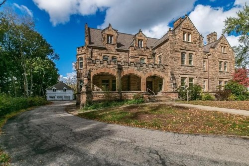 A large, historic stone mansion with intricate architecture, surrounded by trees and a winding driveway.