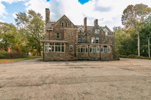 A large, historic stone house with multiple chimneys, surrounded by trees and an empty parking area.