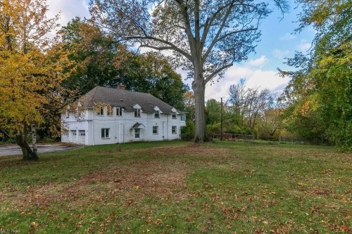 A large white house with a sloped roof, surrounded by trees and a grassy yard, under a partly cloudy sky.