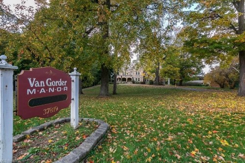 Sign for Van Gorder Manor in a scenic area with autumn leaves and a large historic building in the background.