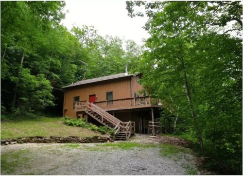 A wooden house surrounded by trees, featuring a staircase and a gravel driveway.