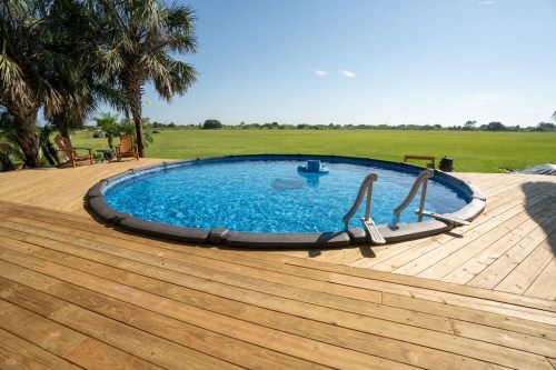 A circular pool with clear blue water on a wooden deck, surrounded by palm trees and open green fields.