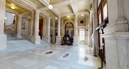Interior view of a grand hall with marble floors, tall columns, and a staircase, featuring ornate decorations and natural light.