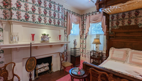 Cozy vintage bedroom with floral wallpaper, a four-poster bed, and elegant furnishings, featuring a fireplace and decorative accents.