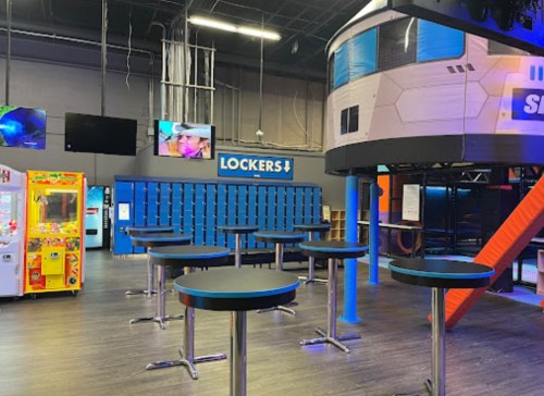 Interior of a recreational facility featuring lockers, arcade games, and tables, with a colorful play structure in the background.