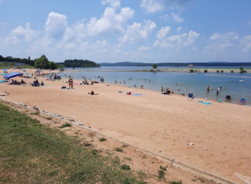 A sandy beach with people swimming and relaxing under a blue sky, surrounded by water and greenery.