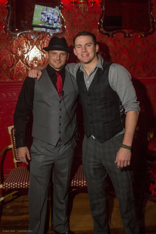 Two men in stylish vests and hats pose together in a vibrant, red-themed venue.
