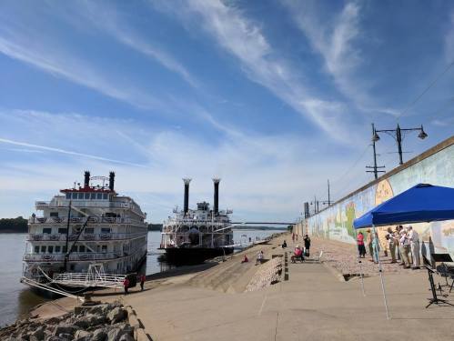 A riverboat docked by a mural-covered wall, with people under a blue tent enjoying the sunny day.