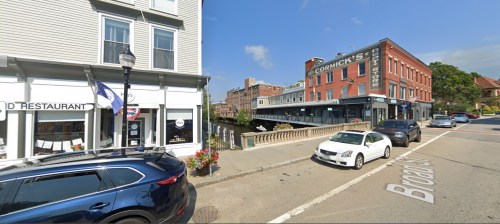 Street view featuring a restaurant, a store, and parked cars along a bridge in a small town.