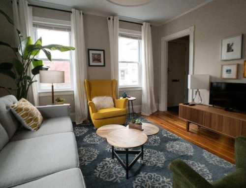 Cozy living room with a gray sofa, yellow armchair, wooden coffee table, and plants, featuring natural light from windows.