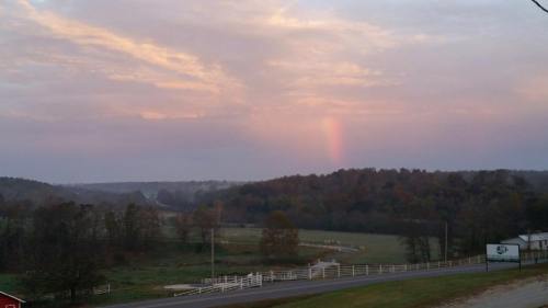 A serene landscape at dusk with a faint rainbow in the sky, surrounded by trees and rolling hills.