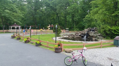 A playground with a pond and waterfall, surrounded by greenery, with a pink bicycle parked nearby.