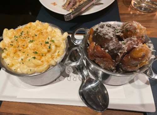 Two metal bowls on a plate: one with creamy macaroni and cheese, the other with roasted potatoes topped with cheese.