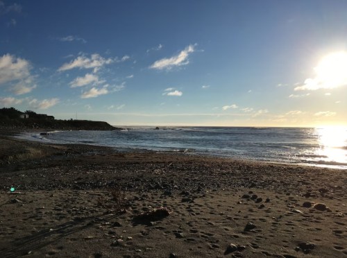 A serene beach scene with gentle waves, pebbles on the shore, and a bright sky with scattered clouds.