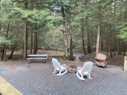 A serene campsite with two white chairs, a fire pit, picnic table, and a grill surrounded by trees and a small stream.
