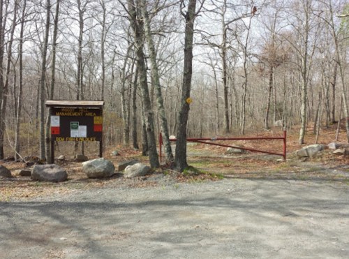 Entrance to a wooded area with a management sign, surrounded by trees and a gravel path. A red gate is partially closed.