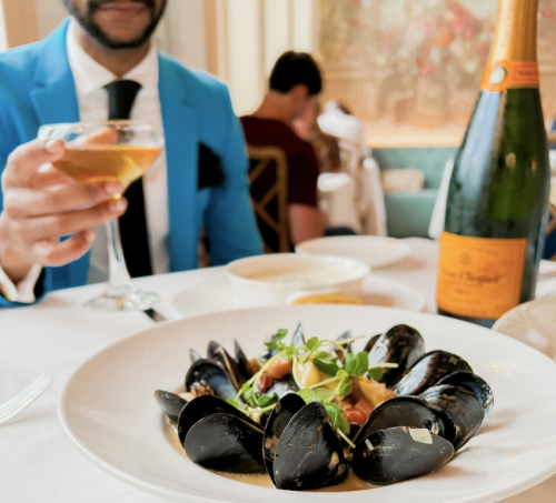 A plate of mussels garnished with herbs, with a cocktail and champagne in the background at a dining table.