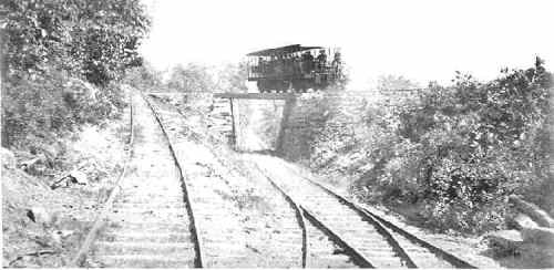 A vintage train on a bridge over a ravine, surrounded by trees and rocky terrain, with parallel tracks leading away.