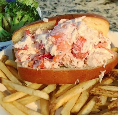 A lobster roll filled with lobster meat on a bun, served with fries and a side of broccoli.