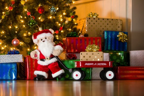 A plush Santa Claus stands beside a red wagon filled with colorful wrapped gifts, in front of a decorated Christmas tree.