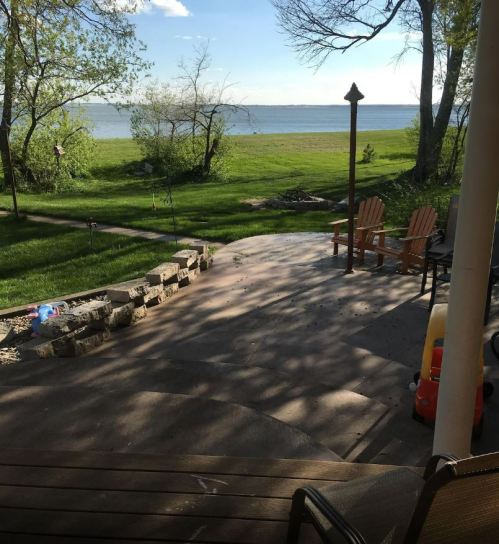 A serene lakeside view from a patio, featuring green grass, trees, and two wooden chairs facing the water.