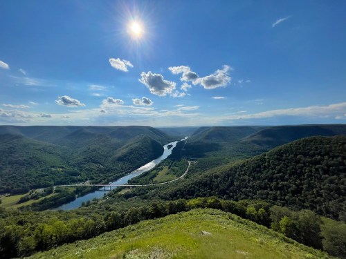 A panoramic view of a winding river surrounded by lush green hills under a bright blue sky with fluffy clouds.