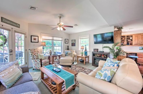 Cozy living room with light-colored furniture, decorative pillows, a ceiling fan, and a TV, surrounded by plants and artwork.