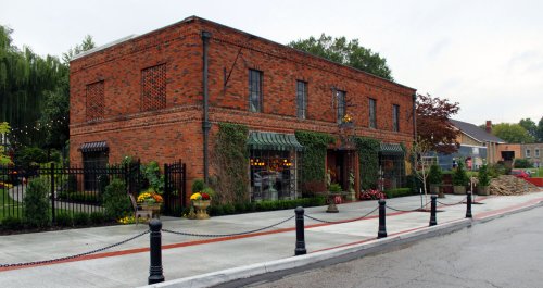 A brick building with green awnings, surrounded by plants and flowers, along a paved street with decorative fencing.