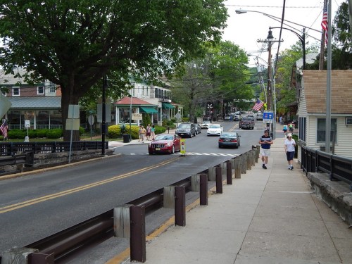 A street scene with pedestrians walking, cars driving, and trees lining the road in a small town.