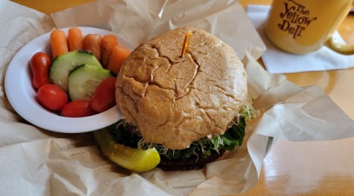 A veggie burger on a plate with lettuce, a pickle, and a side of mixed vegetables including carrots, cucumbers, and tomatoes.
