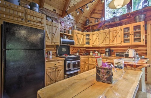Cozy wooden kitchen with black appliances, rustic cabinets, and a large wooden table in a cabin-like setting.