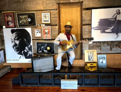 A lifelike statue of a musician playing guitar, surrounded by music memorabilia and photos on a wooden stage.