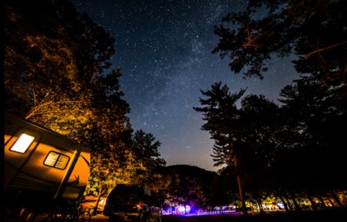 A starry night sky over a campsite, with trees and illuminated tents in the foreground.