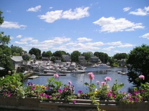 A scenic view of a marina with boats, surrounded by greenery and colorful flowers under a blue sky with fluffy clouds.