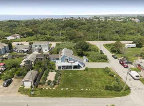 Aerial view of a coastal neighborhood with houses, greenery, and a road leading to the ocean in the background.