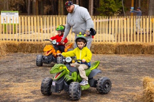 Two children ride toy ATVs shaped like dinosaurs, while an adult supervises in a park with hay bales in the background.